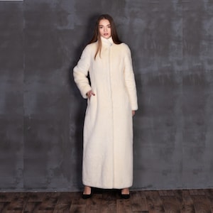 May include: Full-length ivory coat with a high collar and long sleeves. The coat has a button closure and a soft, textured appearance. The model is wearing black heels. The background is a dark gray wall.