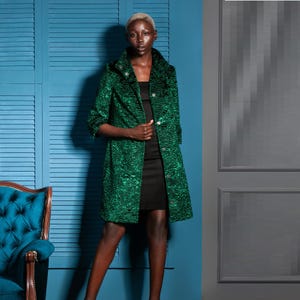 May include: A long, green textured coat with a three-quarter sleeve length. The coat has a button closure and a high collar. The person is wearing a black dress underneath. The background features a blue wall and a teal chair.