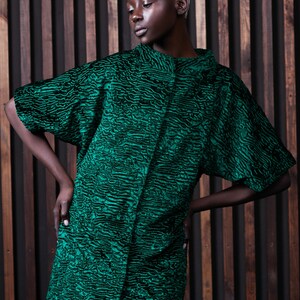 May include: A green textured coat with a black pattern. The coat has short sleeves and a high collar. The model is wearing a black skirt. The background is a wooden wall.