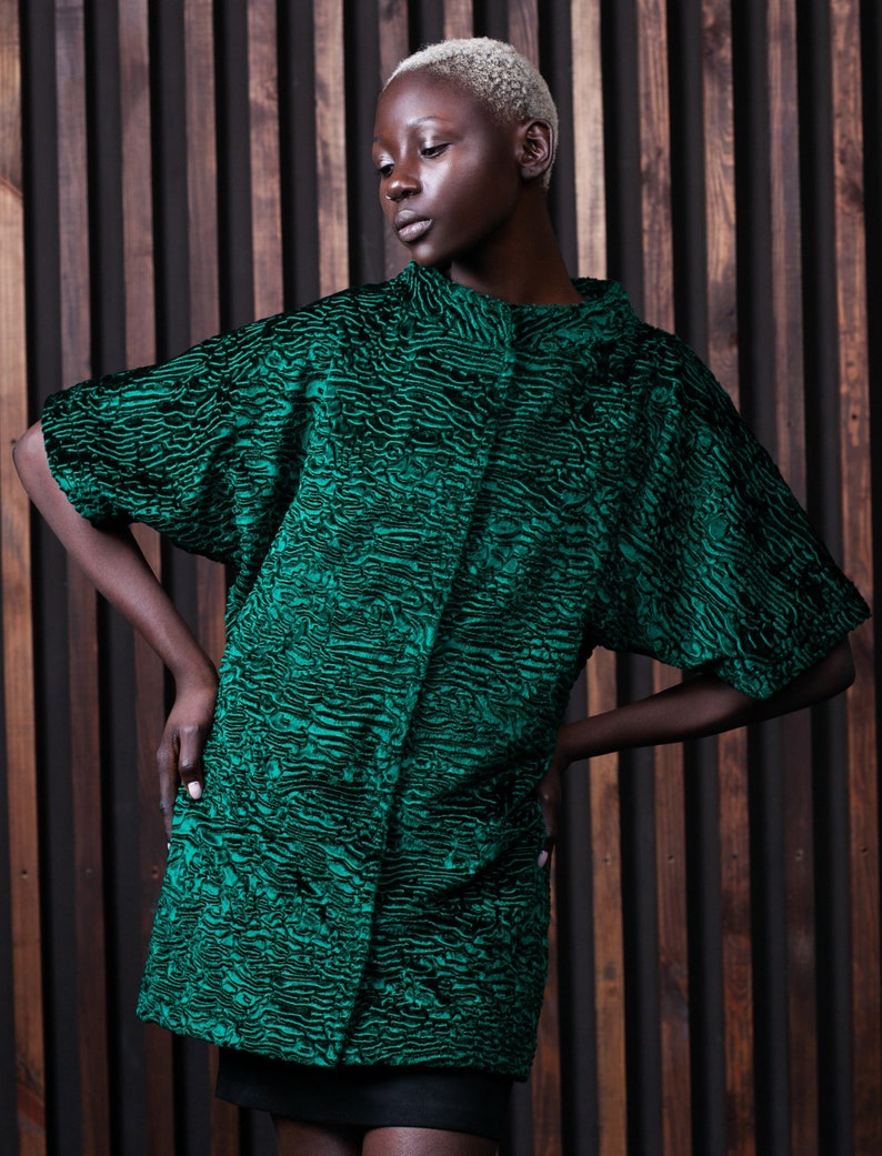 May include: A green textured coat with a black pattern. The coat has short sleeves and a high collar. The model is wearing a black skirt. The background is a wooden wall.