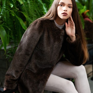 May include: A dark brown faux fur coat with a round collar. The coat is worn over a white top and light grey leggings. The background features lush green foliage.