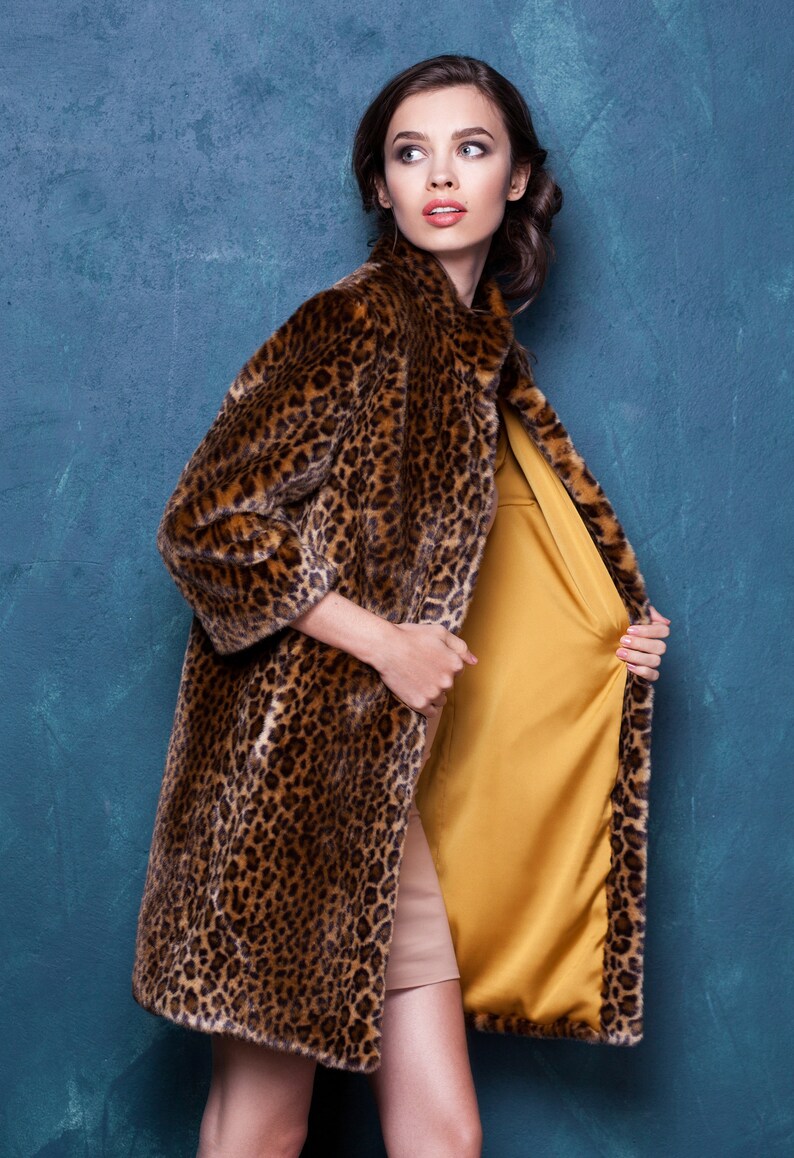 Pu&ograve; includere: Un lungo cappotto leopardato con un motivo marrone e nero. Il cappotto &egrave; aperto, rivelando una fodera in raso giallo senape e un vestito beige. La modella guarda di lato su uno sfondo verde acqua.