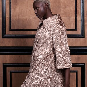 May include: A textured, short-sleeved coat in shades of gold and brown. The coat has a wide collar and a unique, patterned surface. The background is a wooden wall with black trim, creating a contrast with the coat's color.
