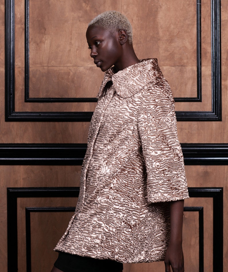 May include: A textured, short-sleeved coat in shades of gold and brown. The coat has a wide collar and a unique, patterned surface. The background is a wooden wall with black trim, creating a contrast with the coat's color.