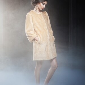 Pu&ograve; includere: Un cappotto in finta pelliccia beige chiaro, lunghezza al ginocchio, con colletto semplice e maniche lunghe. La modella &egrave; in un ambiente fumoso. Il cappotto ha tasche e un'estetica morbida ed eterea. Un capo di abbigliamento classico.