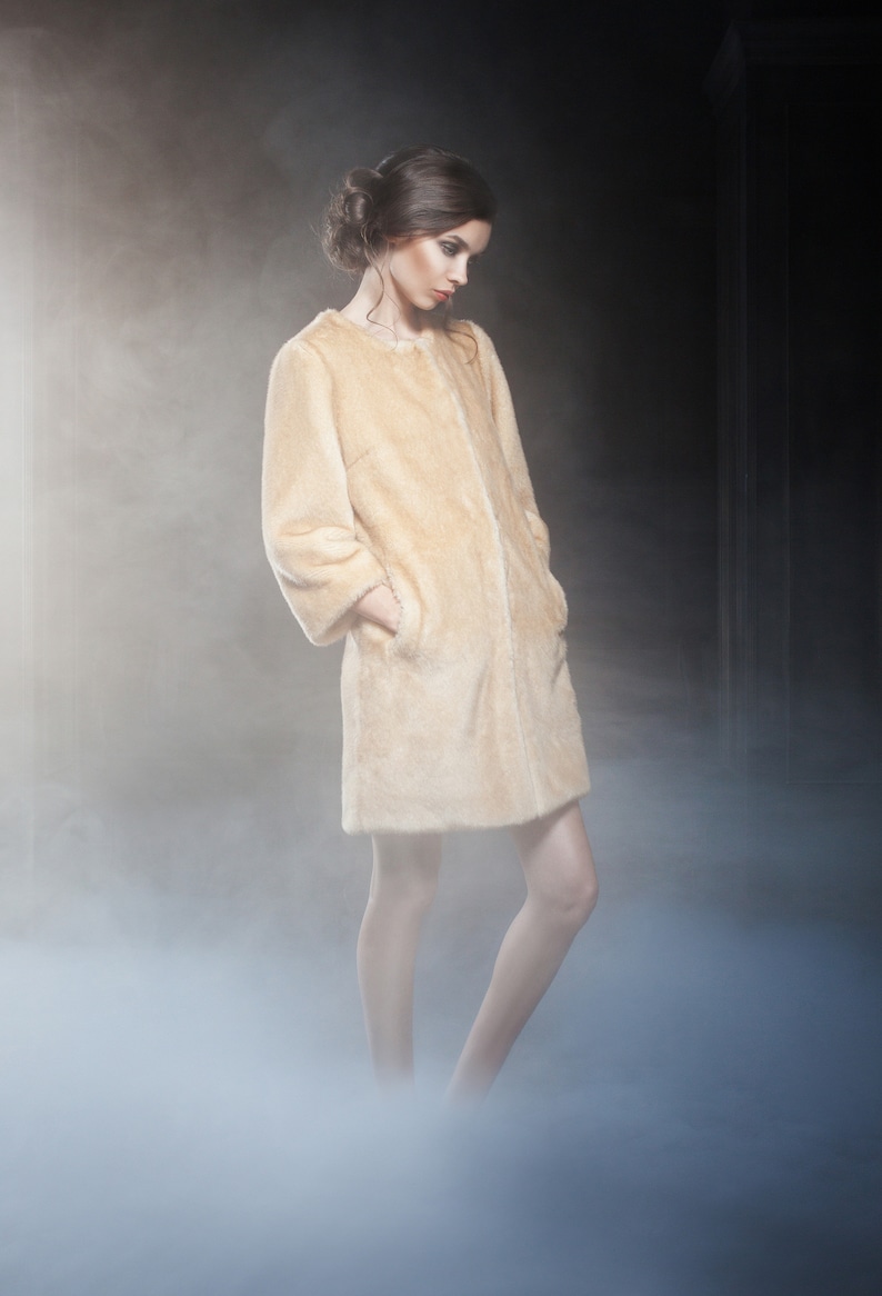 Pu&ograve; includere: Un cappotto in finta pelliccia beige chiaro, lunghezza al ginocchio, con colletto semplice e maniche lunghe. La modella &egrave; in un ambiente fumoso. Il cappotto ha tasche e un'estetica morbida ed eterea. Un capo di abbigliamento classico.