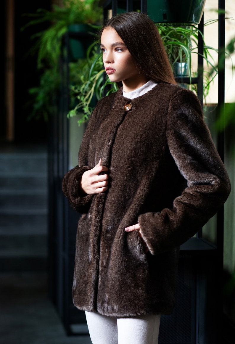 May include: A dark brown faux fur coat with a round collar and a single decorative clasp. The coat has long sleeves and pockets. The model is wearing a white shirt and white trousers. The background includes green plants.
