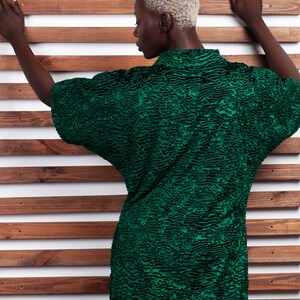 May include: A textured, emerald green top with short sleeves. The top has a unique, patterned design and a high collar. The model is positioned against a wooden slat background.