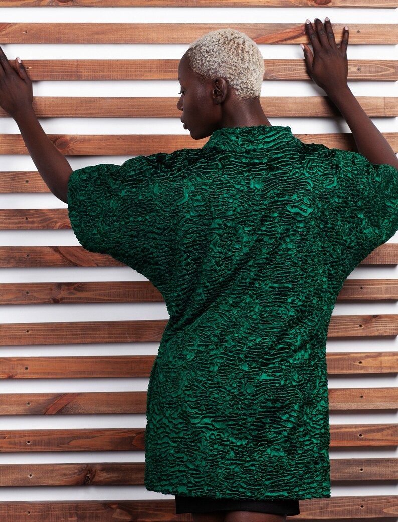 May include: A textured, emerald green top with short sleeves. The top has a unique, patterned design and a high collar. The model is positioned against a wooden slat background.