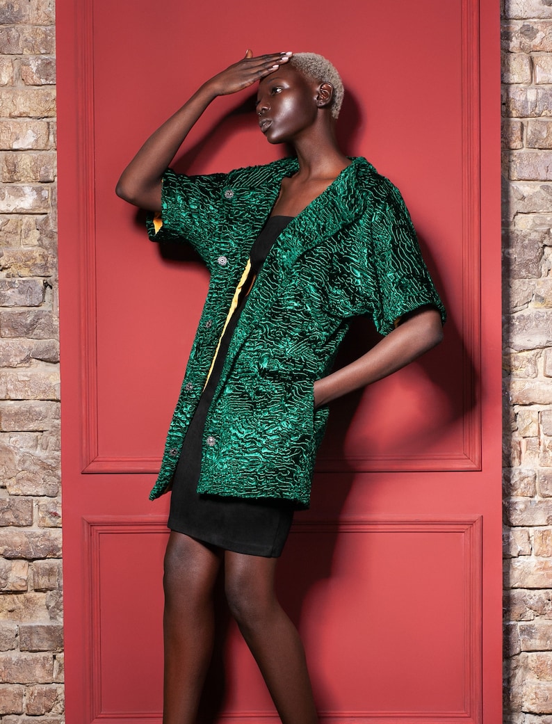 May include: A green textured jacket with short sleeves and a button closure. The jacket has a yellow lining and is worn over a black dress. The background features a red door and a brick wall.