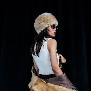 Puede incluir: Una persona con un sombrero de piel sintética beige y gafas de sol, con una camiseta sin mangas blanca y un abrigo de piel sintética marrón. El fondo es una cortina negra lisa.