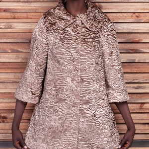 May include: A tan-colored coat with a textured, patterned surface. The coat has a wide collar and three-quarter sleeves. The coat is buttoned up and appears to be knee-length.