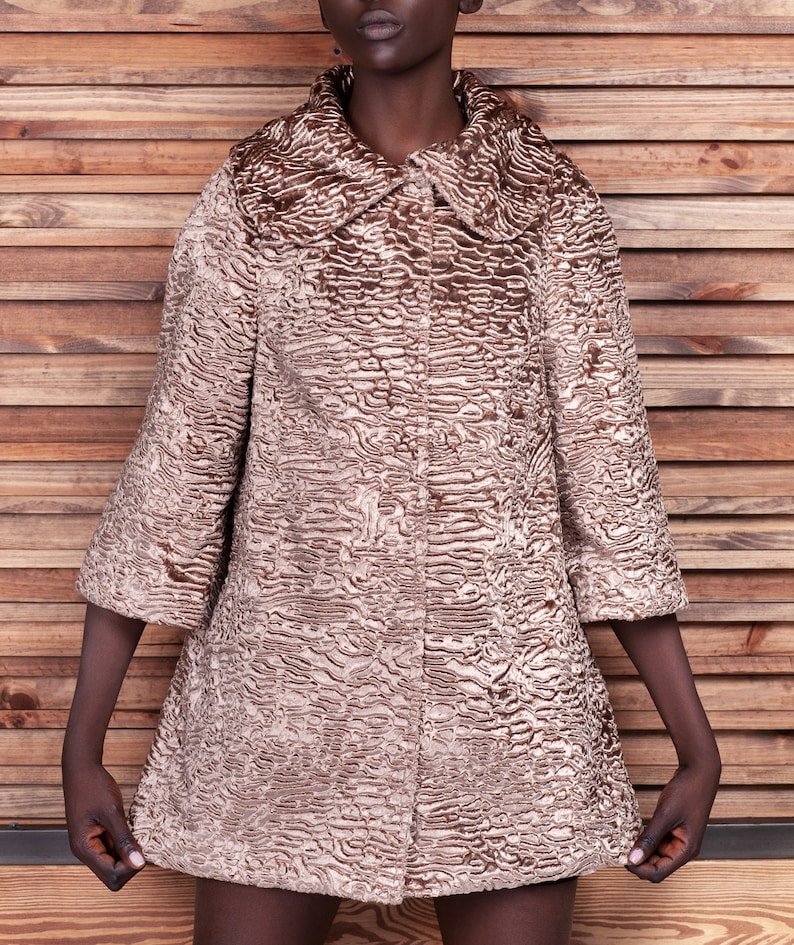 May include: A tan-colored coat with a textured, patterned surface. The coat has a wide collar and three-quarter sleeves. The coat is buttoned up and appears to be knee-length.