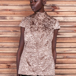 May include: A short-sleeved, high-collared top in a textured, light brown and cream pattern. The garment has a button-up front and a tailored fit. The model is wearing a black skirt, and the background is a wooden wall.