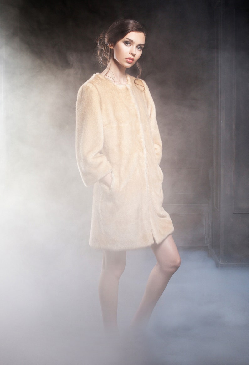 Pu&ograve; includere: Un cappotto in finta pelliccia beige chiaro, con orlo al ginocchio e maniche lunghe. Il cappotto ha un design semplice, senza bottoni. L'immagine &egrave; ambientata in uno studio con uno sfondo fumoso.
