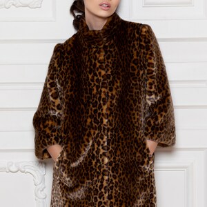 Pu&ograve; includere: Un lungo cappotto in finta pelliccia con stampa leopardata nei toni del marrone e beige. Il cappotto ha un collo alto, maniche a tre quarti e tasche. La modella indossa il cappotto.