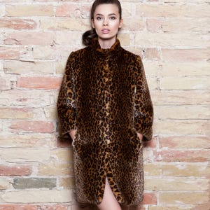 Pu&ograve; includere: Un cappotto lungo con stampa leopardata, collo alto e tasche. Il cappotto presenta un motivo maculato marrone e nero. La modella &egrave; in piedi davanti a un muro di mattoni. Il cappotto sembra essere in ecopelliccia.