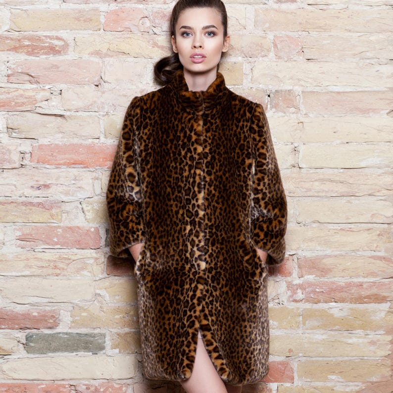 Pu&ograve; includere: Un cappotto lungo con stampa leopardata, collo alto e tasche. Il cappotto presenta un motivo maculato marrone e nero. La modella &egrave; in piedi davanti a un muro di mattoni. Il cappotto sembra essere in ecopelliccia.