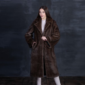 May include: A long, brown faux fur coat with a classic lapel collar. The coat has long sleeves and a straight silhouette, reaching to the ankles. The model is wearing a white top underneath. The coat is a rich, warm brown color.