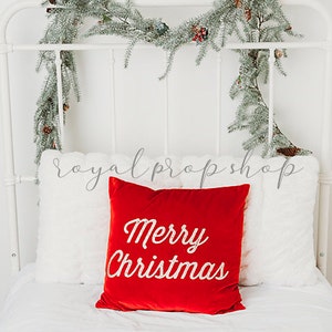 Puede incluir: Una cama con estructura de metal blanco con una almohada de terciopelo rojo que dice "Merry Christmas". La cama está decorada con una guirnalda de ramas de pino.