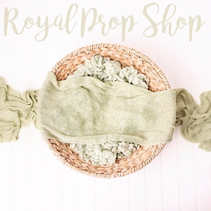Può includere: Un tessuto lavorato a maglia verde chiaro drappeggiato su un cesto intrecciato, con il testo "Royal Prop Shop" sopra il cesto.