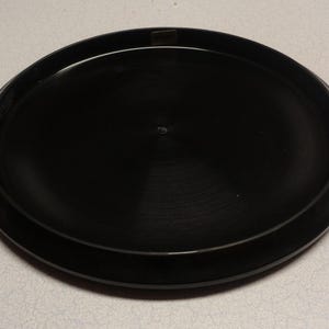 May include: A black disc golf disc with a smooth, circular design. The disc has a slightly raised rim and a flat center. The disc is made of a durable plastic material and is designed for outdoor use. The disc is approximately 8.5 inches in diameter.