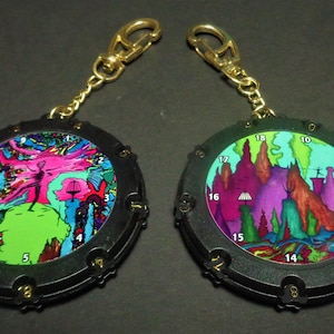 May include: Two black and gold keychains with colorful, surreal landscape art. Each keychain has a round, black frame with gold accents and a gold chain and clasp. The artwork features vibrant colors and numbered details.