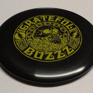 May include: Black disc with the words "Grateful Buzz" in yellow, surrounding a skull and roses design. The disc features a stylized lightning bolt symbol on each side of the skull.