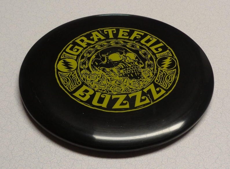 May include: Black disc with the words "Grateful Buzz" in yellow, surrounding a skull and roses design. The disc features a stylized lightning bolt symbol on each side of the skull.