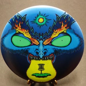 May include: A blue disc golf disc with a monster face design. The monster has green eyes, a yellow and green tongue, and flames on its head. A yellow and green disc golf basket is at the bottom.