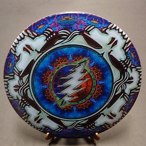 May include: A circular disc featuring a central silver lightning bolt emblem set against a vibrant blue and red mandala-style background. The outer edge displays a repeating white and dark brown abstract pattern with intricate, flowing line work.