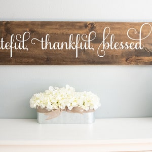 May include: A wooden sign with the text "grateful, thankful, blessed" in white cursive lettering. The sign is hanging on a light blue wall above a white table with a white flower arrangement in a galvanized metal planter and a clear glass jar with white stones and a rope handle.