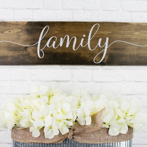 May include: A wooden sign with the word "family" written in white cursive script. The sign is hanging on a white brick wall, and there are white flowers in a metal container below it.