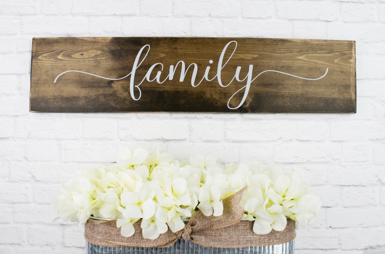 May include: A wooden sign with the word "family" written in white cursive script. The sign is hanging on a white brick wall, and there are white flowers in a metal container below it.