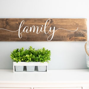 May include: A wooden sign with the word "family" written in white cursive script. The sign is hanging on a white wall above a white dresser with a small potted plant and a glass lantern.