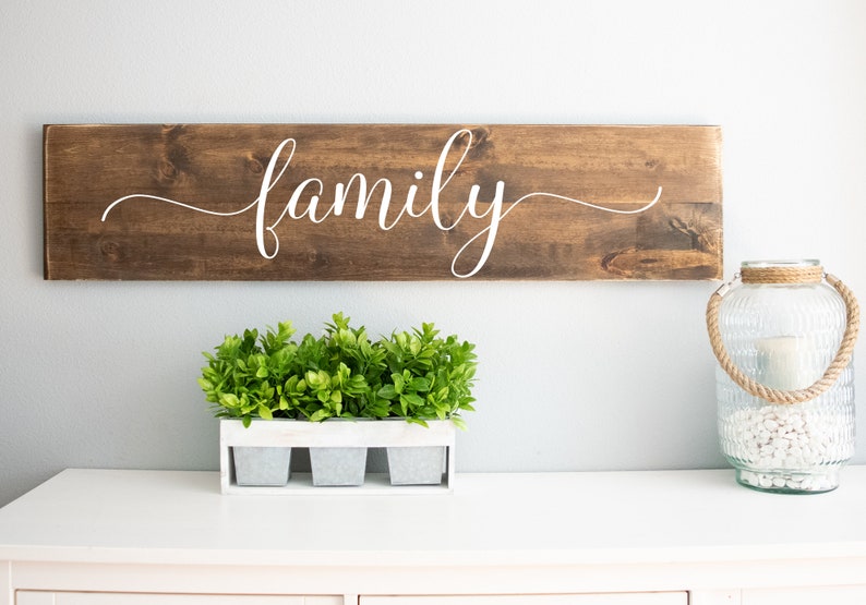 May include: A wooden sign with the word "family" written in white cursive script. The sign is hanging on a white wall above a white dresser with a small potted plant and a glass lantern.