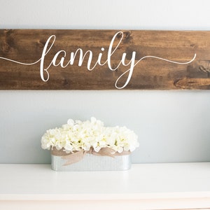 May include: A wooden sign with the word "family" written in white cursive script. The sign is hanging on a light blue wall above a white table with a white flower arrangement in a metal planter and a glass lantern with white stones and a rope handle.