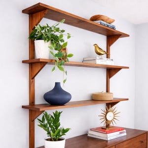 May include: A wooden wall shelf unit with three shelves, displaying decorative items. The shelves hold a white potted plant, a blue vase, a gold bird figurine, books, and a sunburst clock. The unit is made of warm-toned wood.