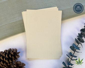 May include: Stack of four cream-colored paper sheets made from 50% European grown hemp. The paper is smooth and has a subtle texture. The paper is on a white surface with a green fabric and a pine cone in the background.
