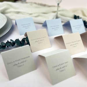 Puede incluir: Tarjetas de sitio plegadas en tonos blancos y azules, dispuestas sobre una mesa. Cada tarjeta muestra nombres y números de mesa en una elegante escritura. Ramas de eucalipto añaden un toque de vegetación. Ideal para bodas.
