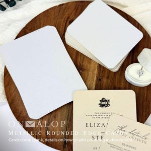 Puede incluir: Una pila de tarjetas rectangulares blancas en blanco con esquinas redondeadas junto a una tarjeta de invitación de boda color crema impresa con una ilustración de cenador negro y el texto The honour of your presence is requested at the wedding.