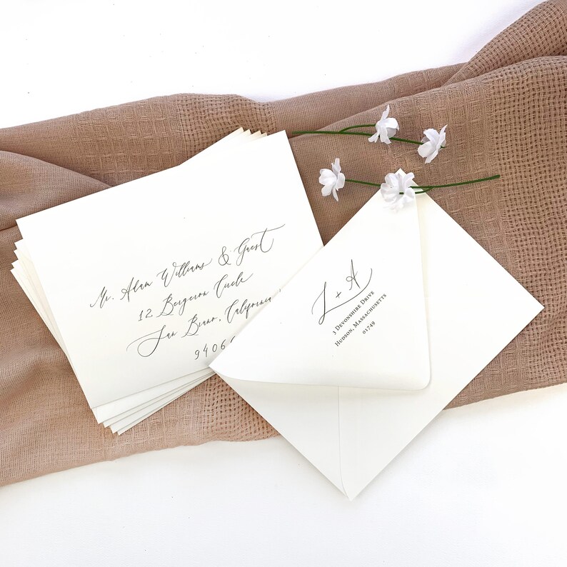 May include: Two white envelopes with calligraphy on them. The envelope on the left has an address written in cursive. The envelope on the right has the initials "L + A" written in cursive and the address "3 Devonshire Drive, Hudson, Massachusetts 01749".