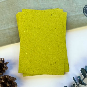 May include: A stack of bright yellow paper sheets with a rough texture. The paper has a subtle speckled pattern and is made from recycled materials. The text on the paper reads "Smells like grass from Bavarian meadows."