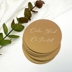 May include: A stack of circular, metallic gold cardstock coasters. The top coaster features white cursive text that reads "Order Blank Or Printed". The coasters have a smooth, matte finish and are arranged in a neat, overlapping vertical pile.
