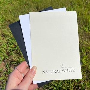 May include: Three sheets of paper, one black, one white, and one off-white with a linen texture. The off-white sheet has the text "linen NATURAL WHITE" printed on it.