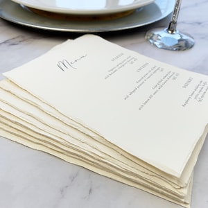 May include: A stack of cream-colored paper menus with a handwritten "Menu" title. The menu items are printed in a simple font. The paper has a rough, deckled edge. A wine glass and a plate are in the background.