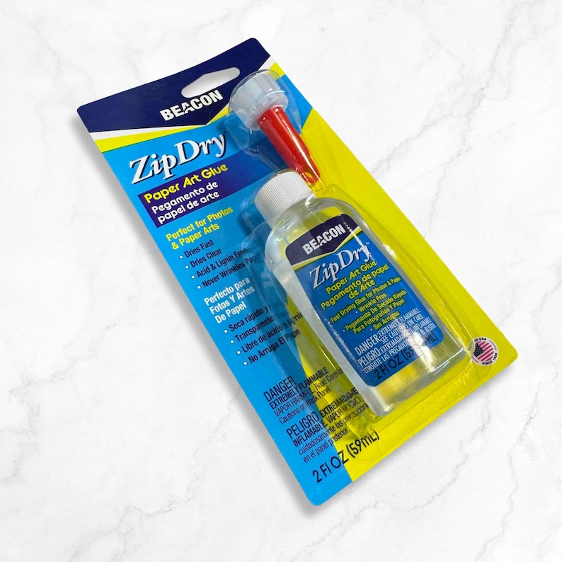 May include: A yellow and blue package containing a clear bottle of Beacon Zip Dry Paper Art Glue. The bottle is 2 fl oz (59 ml) and has a red and white applicator tip. The packaging includes instructions for use and safety warnings.