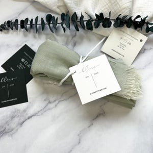 May include: A sage green scarf with frayed edges, tied with a white string and a white tag labeled bliss. Includes two additional square tags, one black and one white, both featuring care instructions and the website address blissboutique.com.