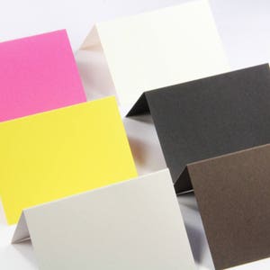 May include: A collection of six folded rectangular cards in various solid colors including bright pink, white, yellow, black, dark brown, and light gray. Each card is positioned upright, displaying a clean, matte texture and a simple fold.