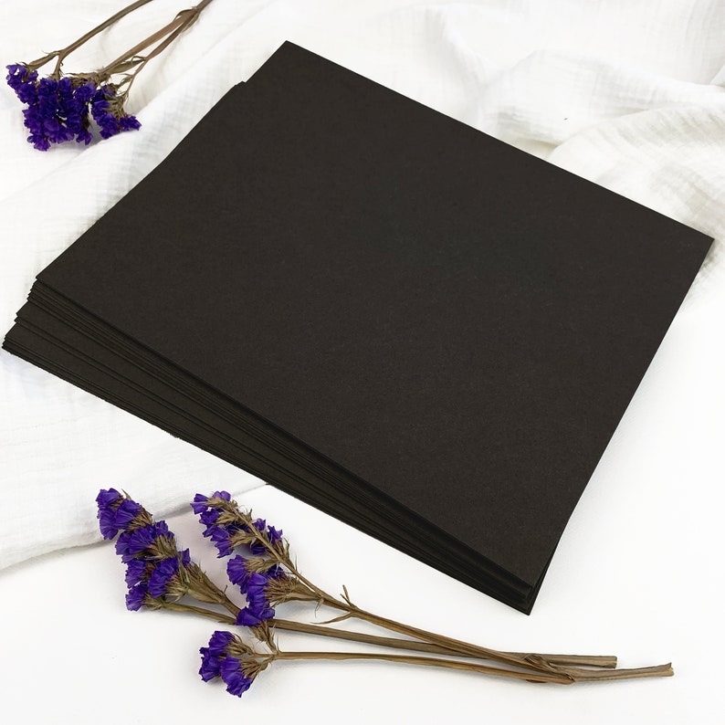 May include: A stack of black paper sheets, likely cardstock or similar, with a textured surface. The paper is arranged on a white cloth, with sprigs of purple dried flowers adding a touch of color.